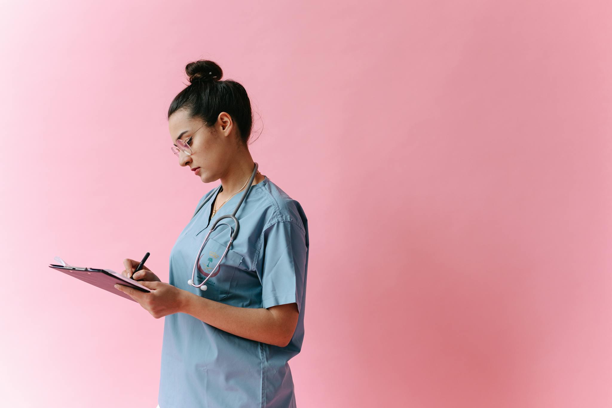 Female nurse in scrubs writing on a clipboard with a stethoscope on a pink background.